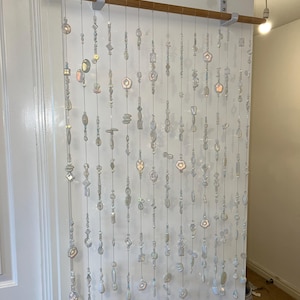 May include: A decorative beaded curtain featuring iridescent beads in various shapes and sizes. The beads are strung on clear cords and suspended from a wooden rod. The curtain hangs against a white wall, adding a touch of elegance to the space.