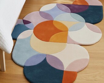 May include: Three colorful, abstract rugs with a geometric design. The rugs feature a combination of circles in shades of blue, orange, purple, and cream. The rugs are placed on a wooden floor near a bed with a white comforter and a purple pillow.