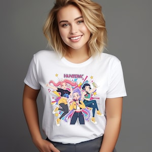 Puede incluir: Camiseta blanca con un gráfico colorido de tres personajes de estilo anime y el texto "HUNTXX". Los personajes están en poses dinámicas con colores vibrantes, sobre un fondo de estrellas y formas geométricas. La camiseta es de cuello redondo.