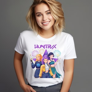 Puede incluir: Camiseta blanca con la palabra "HUNTR/X" en morado y un gráfico de tres mujeres de estilo anime frente a una estrella y fuegos artificiales. Las mujeres visten diferentes atuendos, incluyendo una chaqueta amarilla y un vestido turquesa.