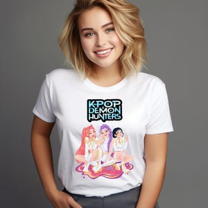Può includere: T-shirt bianca con la scritta "KPOP DEMON HUNTERS" sopra una grafica a cartoni animati di tre figure. Le figure hanno diversi colori di capelli e indossano abiti bianchi con dettagli dorati. Lo sfondo è grigio.