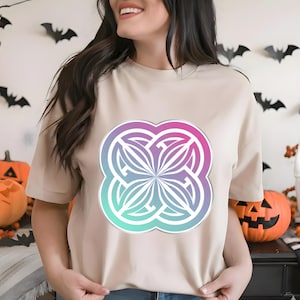 Peut inclure: Un t-shirt beige avec un grand motif coloré. Le motif est un design symétrique stylisé avec un dégradé de rose, de violet et de sarcelle. Le t-shirt est à manches courtes et a un col rond. Des décorations d'Halloween sont en arrière-plan.