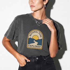 Peut inclure: Un t-shirt gris anthracite à manches courtes avec une impression graphique représentant un soleil jaune se levant sur des montagnes bleues et un chemin sinueux. Le texte "NATIONAL PARK EXPLORER" est imprimé en noir dans une bordure arquée.