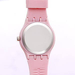 May include: A light pink wristwatch with a matching strap. The watch has a round face with a silver-colored back and a pink band. The watch band has a series of holes for adjustment.