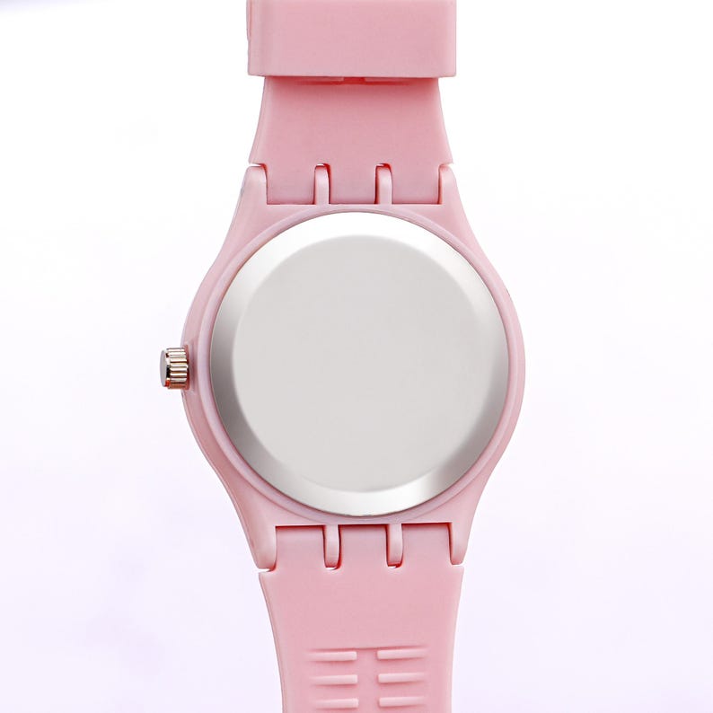 May include: A light pink wristwatch with a matching strap. The watch has a round face with a silver-colored back and a pink band. The watch band has a series of holes for adjustment.