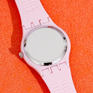 May include: A light pink wristwatch with a silver-colored bezel and a white watch face. The watch band is made of a matching light pink material. The watch is displayed against an orange background.