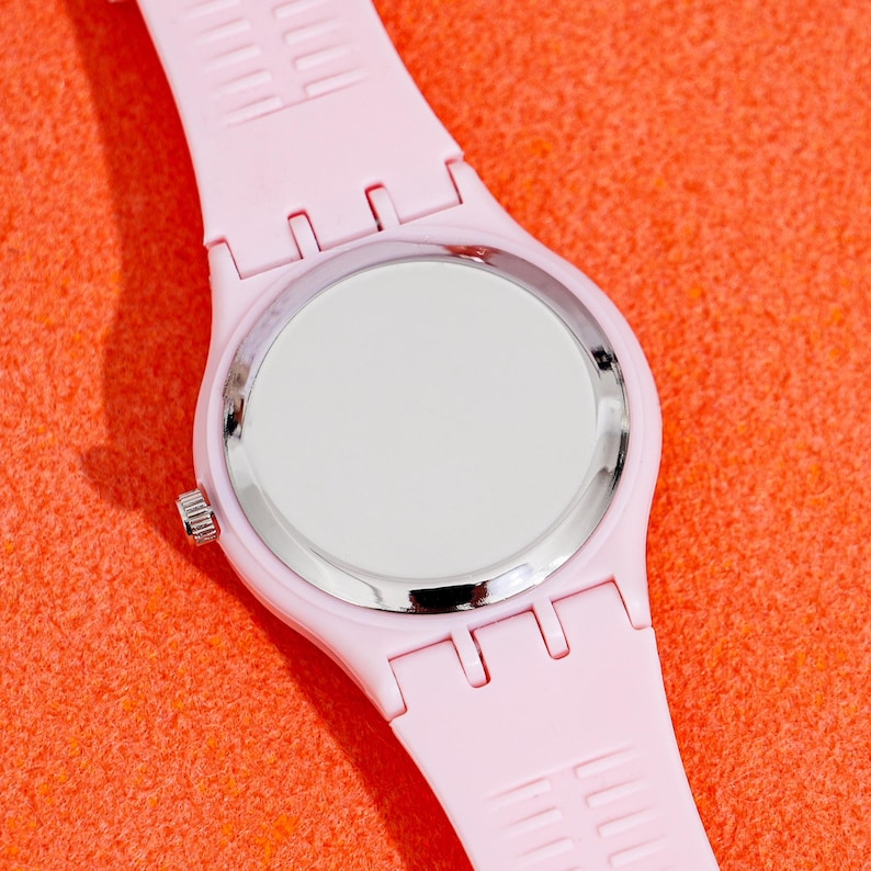 May include: A light pink wristwatch with a silver-colored bezel and a white watch face. The watch band is made of a matching light pink material. The watch is displayed against an orange background.