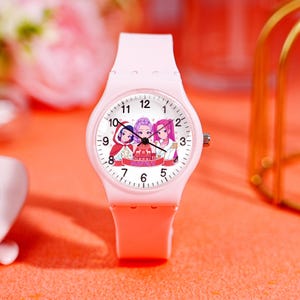 May include: A light pink wristwatch with a round face featuring an illustration of three cartoon characters. The watch has a matching pink band and a silver crown. The watch face displays the numbers 1 through 12 and the text "FUNTOX".