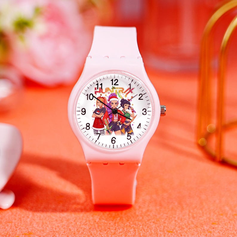 May include: A light pink wristwatch with a white face featuring a colorful graphic of three characters. The watch has a matching pink band and a silver-colored crown. The word "HUNTRIX" is printed on the watch face.