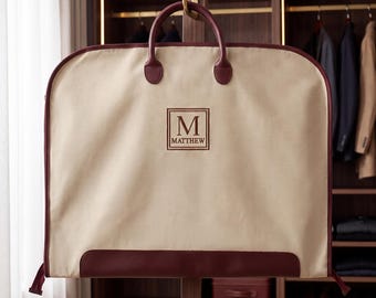 May include: A beige garment bag with dark red trim and handles. The center features a square embroidered patch with the letter M and the name MATTHEW printed below it. The bag is constructed with a sturdy frame and a smooth, fabric exterior.