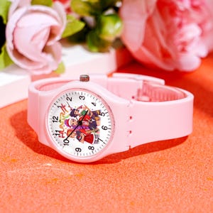 May include: A light pink wristwatch with a white watch face featuring a colorful cartoon design. The watch has a matching pink band and a silver-colored crown. The watch face displays the numbers 1 through 12.