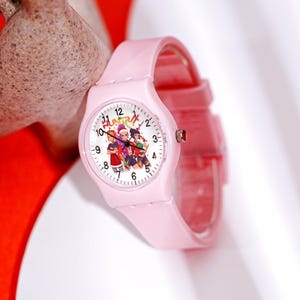May include: A pink wristwatch with a round face featuring a colorful graphic of three cartoon characters. The watch has a pink band and a silver crown. The watch face displays the numbers 1 through 12.