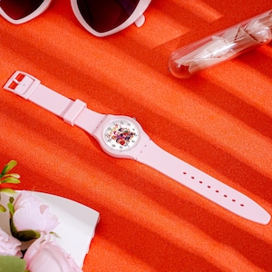 May include: A light pink wristwatch with a floral design on the face. The watch has a matching pink strap and a buckle closure. The watch is set against an orange background, with heart-shaped sunglasses and a glass tube visible.