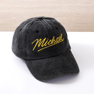 Può includere: Un cappellino da baseball nero con il nome "Michael" ricamato in giallo. Il cappellino ha una visiera curva e un aspetto leggermente sbiadito.