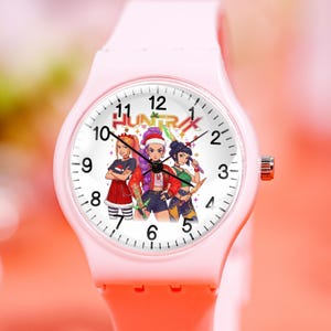 May include: Pink wristwatch with a white face featuring three cartoon characters and the word "HUNTRAX". The watch has a pink band and a silver-colored crown. The watch face displays black numerals and hands.