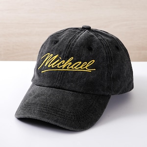 Puede incluir: Gorra de béisbol negra con el nombre "Michael" bordado en escritura amarilla. La gorra tiene una visera curva y una apariencia ligeramente desgastada, lo que sugiere un estilo vintage. El sombrero está hecho de tela suave.