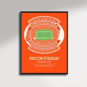 May include: A framed print featuring a stylized top-down view of Paycor Stadium in Cincinnati, Ohio. The stadium is depicted in white lines against a vibrant orange background, with a green field at the center. Text includes the stadium name and coordinates.