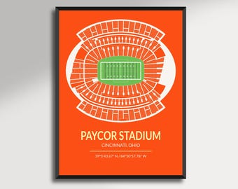 May include: A framed print featuring a stylized top-down view of Paycor Stadium in Cincinnati, Ohio. The stadium is depicted in white lines against a vibrant orange background, with a green field at the center. Text includes the stadium name and coordinates.