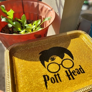 以下が含まれることがあります： 装飾的な縁取りのある、長方形の金色のグリッタートレイ。 トレイには、メガネをかけたキャラクターの顔の黒いシルエットと、様式化されたフォントで「Pott Head」という言葉が描かれています。 緑の植物が入ったテラコッタの鉢が背景にあります。