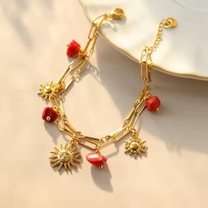 Peut inclure: Bracelet doré avec un design à maillons, des perles de corail rouge et des breloques en forme de soleil. Le bracelet est présenté sur une surface blanche, mettant en valeur ses détails et sa palette de couleurs chaudes.