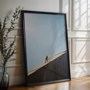 May include: A framed art print featuring a person walking on a rooftop against a blue sky. The black frame leans against a white wall, next to a glass vase with branches. The artwork is displayed on a wooden floor.