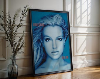 May include: Framed poster with a blue-toned portrait of a woman with blonde hair. The poster has the text "Britney Spears" in pink at the top and "In The Zone" at the bottom. The poster is leaning against a white wall and wooden floor.