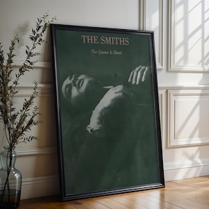 May include: Framed poster featuring the album art for The Smiths' "The Queen Is Dead." The poster has a black frame and a dark green background. The album title and band name are in a light pink font. The image shows a black and white photo of a person.
