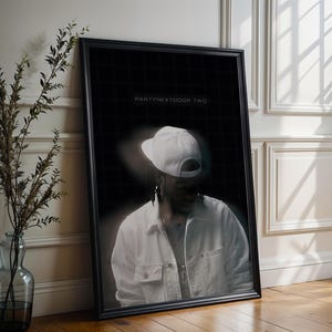 Puede incluir: Póster enmarcado en negro con una persona que lleva una gorra y una chaqueta blancas. El texto "PARTYNEXTDOOR TWO" se muestra en la parte superior del póster. El póster está apoyado contra una pared blanca y un suelo de madera.