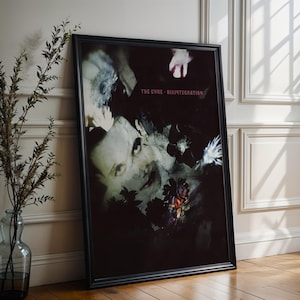 May include: Framed poster featuring the album art for The Cure's Disintegration. The artwork is dark with a portrait of a person and floral elements. The poster is in a black frame and leans against a white wall and wooden floor.