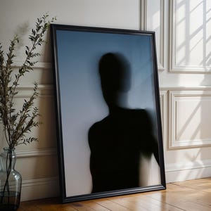 May include: A framed art print featuring a silhouette of a person against a gradient blue background. The black frame leans against a white wall with decorative molding. A glass vase with branches sits nearby.