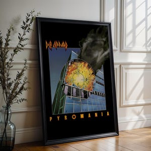 Könnte beinhalten: Gerahmtes Poster mit dem Albumcover von Def Leppard's Pyromania. Das Kunstwerk zeigt ein Gebäude mit einem Ziel über einer Explosion. Das Poster ist in einem schwarzen Rahmen und lehnt an einer weißen Wand. Musik-Kunstwerk.