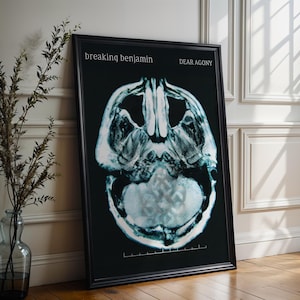 May include: Framed art print showing a grayscale image of a human skull, similar to an X-ray. The print is titled "DEAR AGONY" by "breaking benjamin" in white text on a black background. The artwork is displayed on a wooden floor.