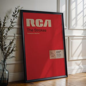Puede incluir: Un póster enmarcado con fondo rojo y marco negro. El póster presenta el texto "RCA" en negrita, en color blanquecino, con "The Strokes" y "Comedown Machine" debajo. Una pequeña etiqueta está adherida en la esquina inferior derecha.