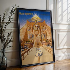 May include: Framed poster featuring the Iron Maiden band logo and artwork of an Egyptian-themed scene with a pyramid and figures. The poster has a black frame and is leaning against a wall.