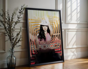 May include: Framed poster of Britney Spears, with the text "Britney Spears Blackout". The poster is in a black frame, leaning against a white wall. The image uses pink, orange, and black in its design.