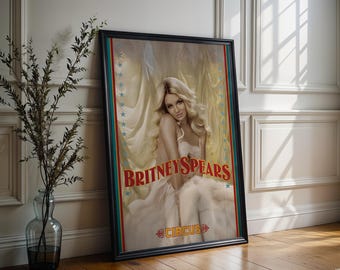 May include: Framed poster of Britney Spears in a white dress, with the text "BRITNEY SPEARS" in red. The poster has a circus theme with a light yellow background and a black frame. The word "CIRCUS" is at the bottom.