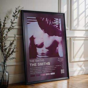 May include: Framed poster featuring a monochromatic image of a person's torso. The poster has the band name "THE SMITHS" in white text, along with song titles and a QR code. The frame is black, and the poster leans against a white wall.