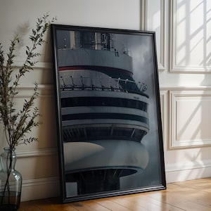 Op de afbeelding: Ingelijste print van een hoge, cilindrische structuur met een wit en grijs exterieur, met een rode streep. De print leunt tegen een witte muur en houten vloer, met een vaas met gedroogde planten.