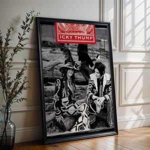 May include: Framed black and white photograph featuring two people in ornate black and white outfits, seated on a log. A red banner with the text "ICKY THUMP" is displayed above the photograph. The frame is black, and the image is leaning against a white wall.