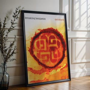 May include: Framed poster with the band name "Breaking Benjamin" and album title "Saturate". The artwork features a circular design in red and orange hues against a yellow backdrop. The poster is presented in a black frame.
