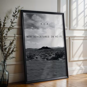 May include: Black and white framed poster featuring a desert landscape under a cloudy sky. The text "R.E.M. NEW ADVENTURES IN HI-FI" is displayed in a minimalist font. The poster is leaning against a white wall next to a vase with branches.