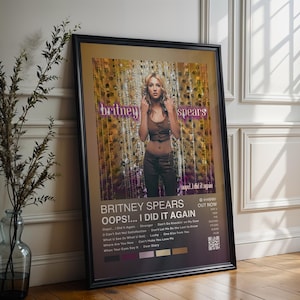 May include: Framed poster of Britney Spears' "Oops!... I Did It Again" album. The poster shows Spears in a brown top and trousers, with the album title and tracklist. The poster is in a black frame and leans against a white wall, with a vase of foliage.