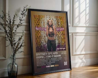 May include: Framed poster of Britney Spears' "Oops!... I Did It Again" album. The poster shows Spears in a brown top and trousers, with the album title and tracklist. The poster is in a black frame and leans against a white wall, with a vase of foliage.