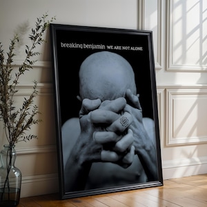 May include: A framed black and white print showing a bald figure with hands clasped in front of the face. The print has the text "breaking benjamin we are not alone" at the top. The frame is black and leans against a white wall.