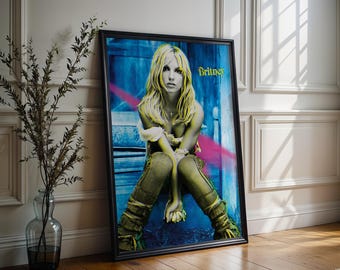 May include: Framed poster featuring a portrait of a blonde woman with the name "Britney" in gold lettering. The image has a blue and pink background, with the subject wearing a white top and green trousers. The poster is in a black frame.