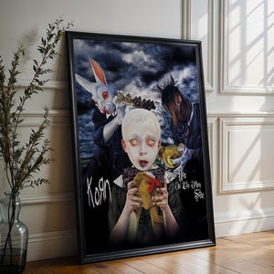 Può includere: Stampa artistica incorniciata raffigurante un bambino con occhi luminosi che tiene un orsacchiotto, circondato da figure in una scena oscura e surreale. L'opera d'arte include un coniglio e una figura simile a un cavallo, con il nome della band "Korn" e la frase "You on the Other Side".