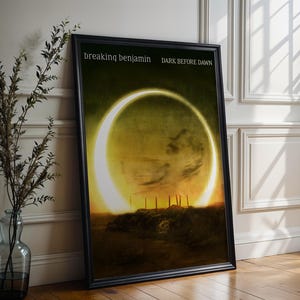 May include: A framed poster of the "Dark Before Dawn" album art by Breaking Benjamin. The image shows a bright, yellow-orange ring against a dark, textured backdrop. The poster, in a black frame, leans against a white wall, with the text "Dark Before Dawn".