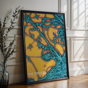 以下が含まれることがあります： 黄色、オレンジ、ティール色の抽象的なデザインの額入りアートプリント。アートワークは黒いフレームで、「THE STROKES」の文字が白で書かれています。プリントは白い壁に立てかけられています。