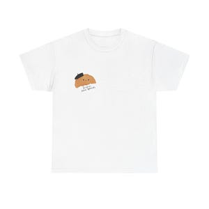 Könnte beinhalten: Ein weißes T-Shirt mit kurzen Ärmeln und einer kleinen Grafik auf der linken Brustseite. Die Grafik zeigt ein hellbraunes Croissant mit einer schwarzen Baskenmütze sowie den kursiven Text "Bonjour mon amour" in schwarzer Schrift darunter.