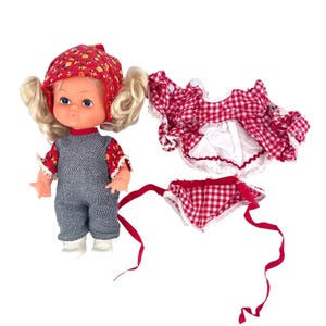 May include: A vintage doll with blonde pigtails, wearing a red floral bonnet, a red floral and denim jumpsuit, and white shoes. A red and white checkered dress and bonnet with red ribbons are next to the doll.